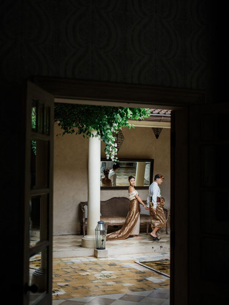 Bali couple photoshoot at Balquisse Heritage Hotel with couple in traditional Balinese outfit walking through an intimate heritage courtyard framed by doorway