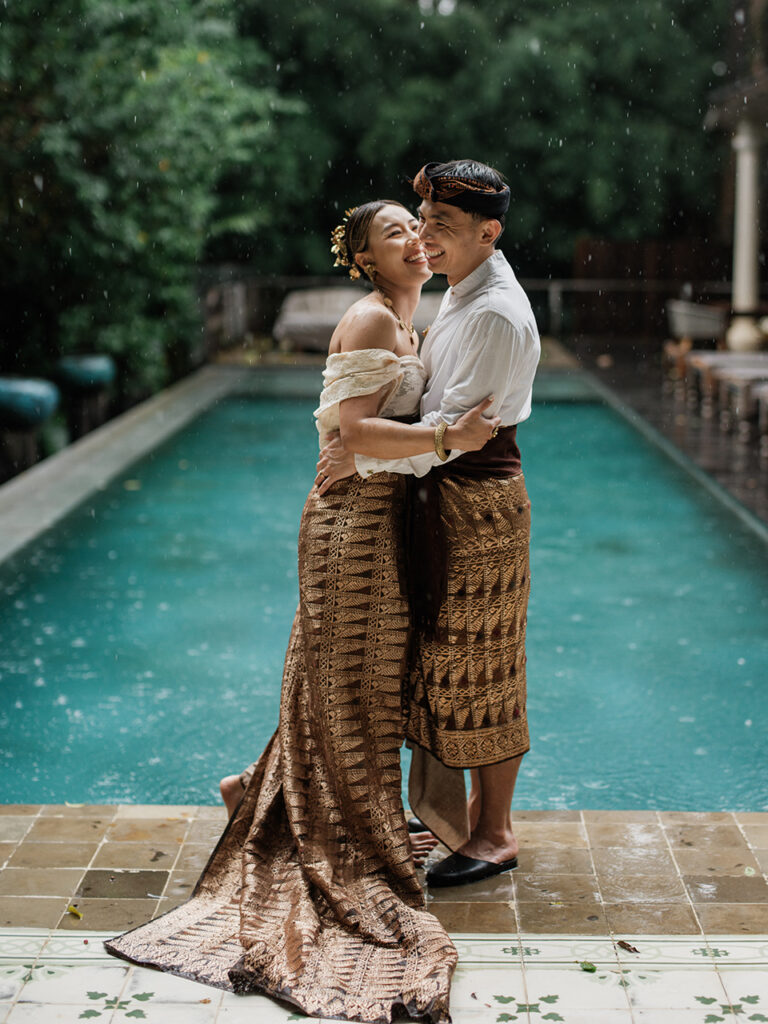 Bali couple photoshoot at Balquisse Heritage Hotel with couple embracing in traditional Balinese outfit by the pool in soft rain