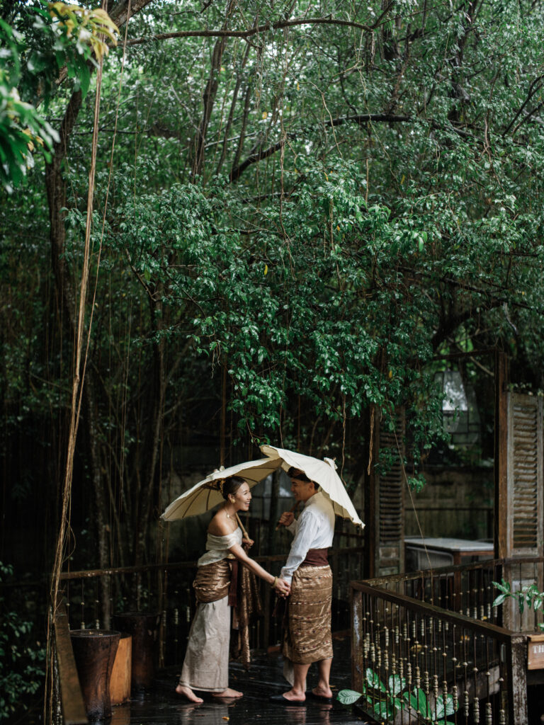 Bride and groom walking under umbrellas during a rainy couple session at Balquisse Heritage Hotel Jimbaran Bali surrounded by tropical greenery.