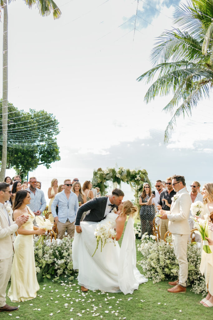 Villa Pemutih wedding Bali ceremony moment with bride and groom kissing, guests celebrating and white floral setup in Uluwatu cliffside venue