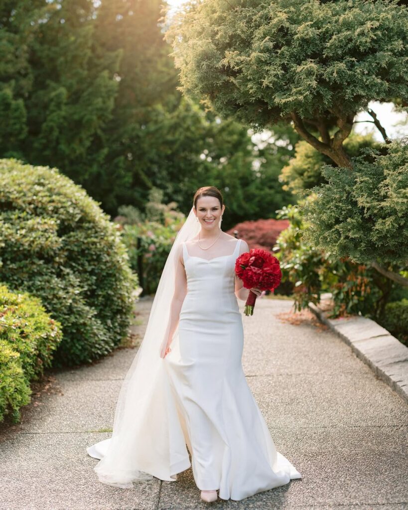 Amanda walking through Cecil Green Park House gardens in white wedding gown with red bouquet, Vancouver
