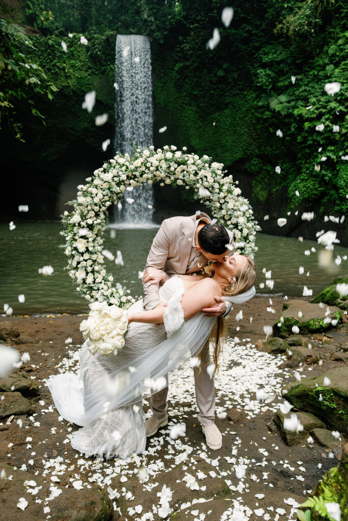 Tibumana Waterfall elopement in Bali with couple kiss under floral arch and falling petals