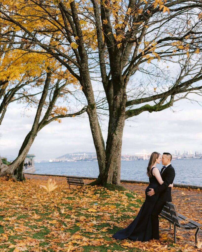 Amanda and Tony standing close under autumn trees at Stanley Park waterfront, Vancouver skyline in distance