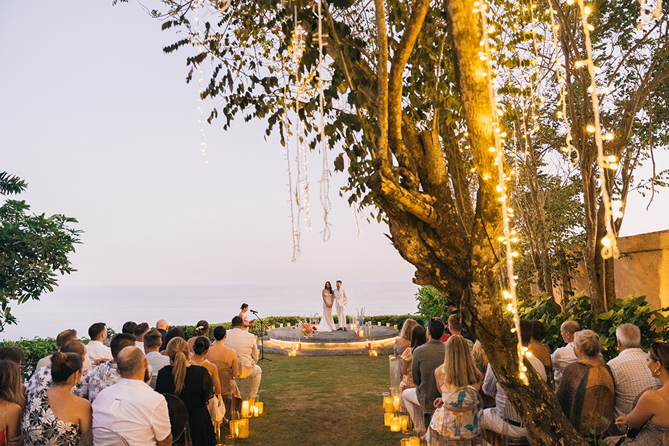 Intimate sunset wedding Wonderland Uluwatu ceremony with guests seated facing a candlelit stage and ocean view at dusk