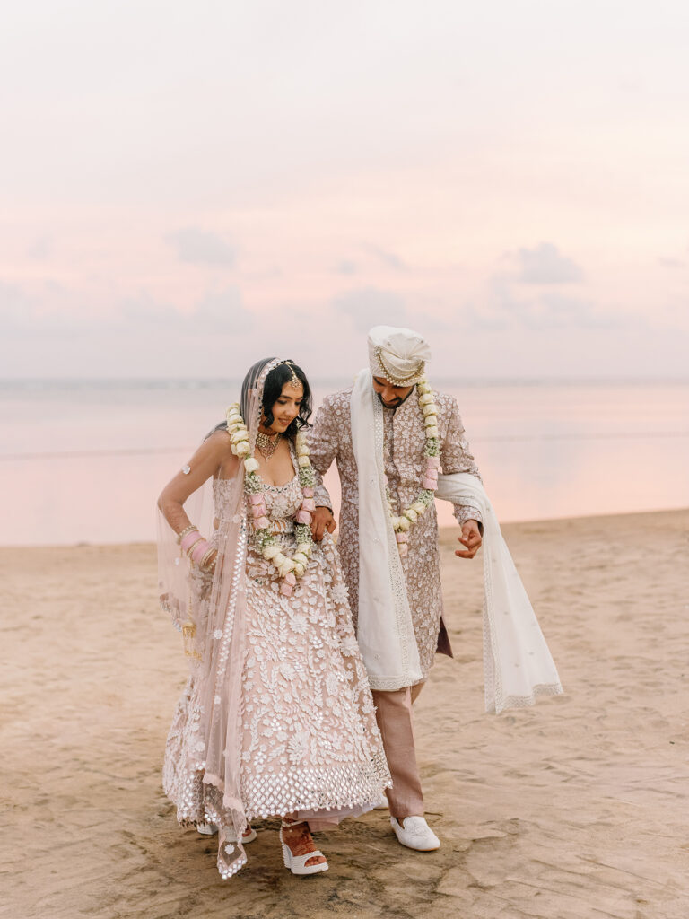 Indian bride and groom walking on Bali beach at sunset — traditional lehenga and sherwani against soft pink sky