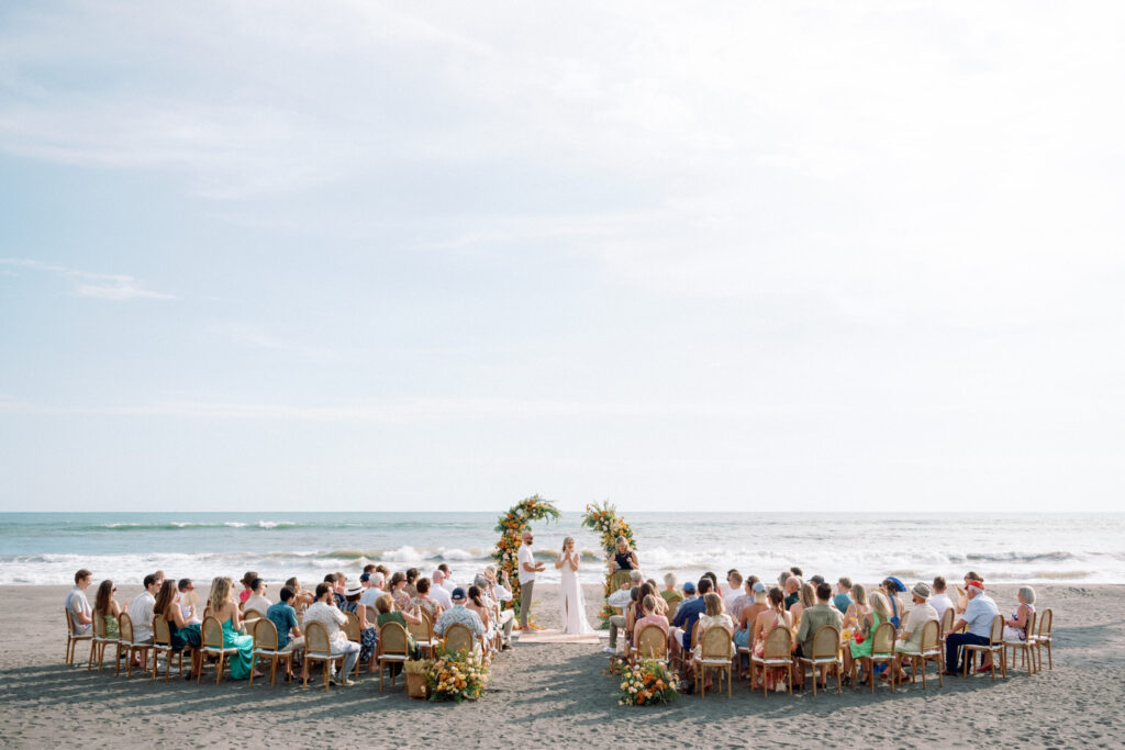 intimate elegant beach wedding ceremony in Bali at Villa Vedas with ocean backdrop, floral arches, and guests seated around the couple