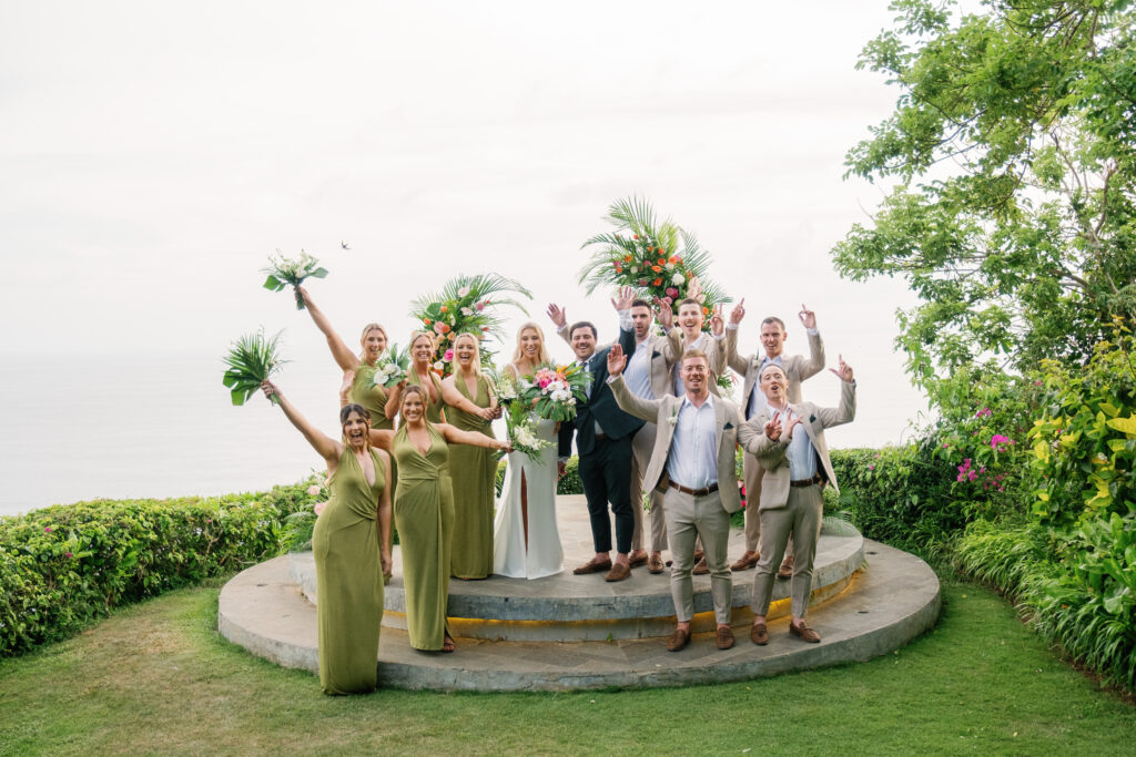 Wonderland Uluwatu wedding group photo with bride, groom, and bridal party celebrating on a cliff overlooking the ocean in Bali