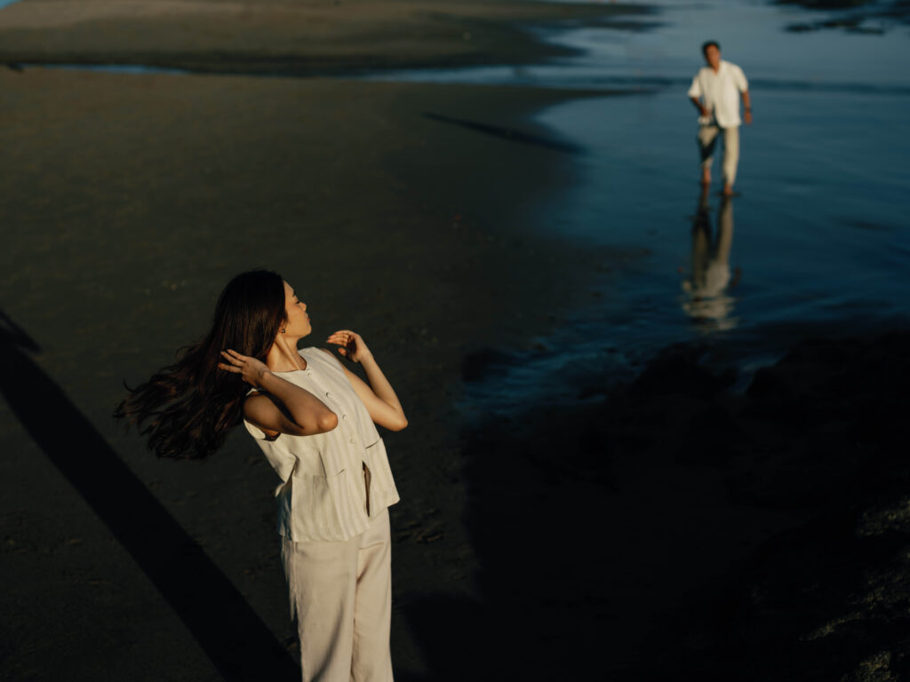 Honeymoon photoshoot Bali candid moment at Echo Beach with bride adjusting her hair in warm sunlight while groom walks in the distance along the shoreline, creating a natural storytelling composition.