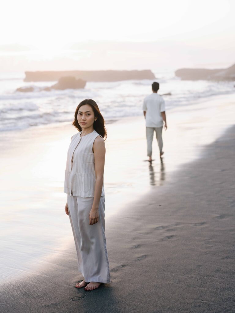 Honeymoon photoshoot Bali at Echo Beach featuring a bride standing barefoot on black sand during soft sunset light while her partner walks along the shoreline in the background, creating a calm and cinematic long-distance connection.