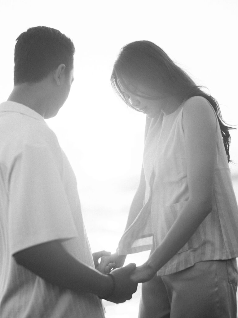 Black and white honeymoon photoshoot in Bali capturing an intimate moment at Echo Beach as a couple gently holds hands in soft backlight, creating a quiet and emotional connection.