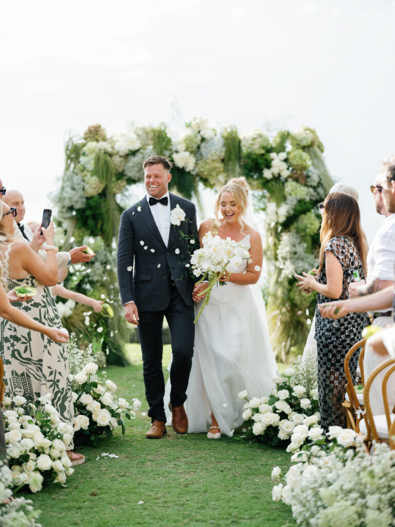 Bride and groom walking down the aisle after their oceanfront wedding ceremony at Villa Pemutih Bali surrounded by white floral arrangements and cheering guests.