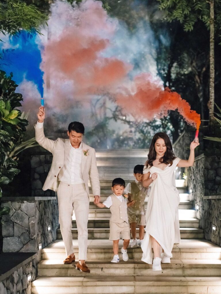 Terry and Jimmy entering wedding reception with their two sons holding smoke bombs at Padma Resort Ubud — family entrance night Bali wedding