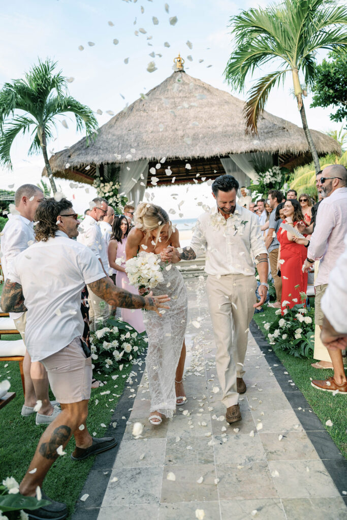 Bride and groom walking back down the aisle at a tropical Bali wedding ceremony while guests throw white flower petals around them at Nusa Dua Beach Hotel gazebo.