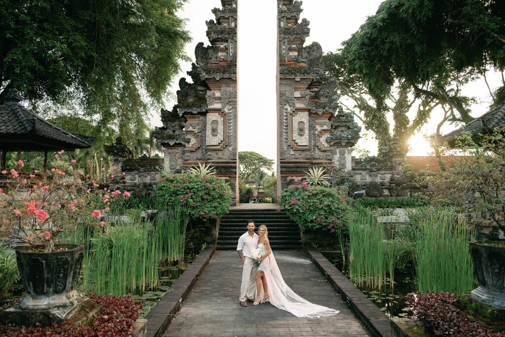 Bride and groom standing beneath a towering traditional Balinese temple gate at Nusa Dua Beach Hotel in Bali, surrounded by tropical gardens, reflective ponds, and warm golden hour light.