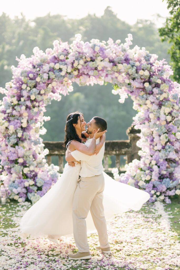 Micaela and Luis lifting each other in front of purple floral arch at Puri Wulandari wedding Ubud Bali — Luxima Wedding photography