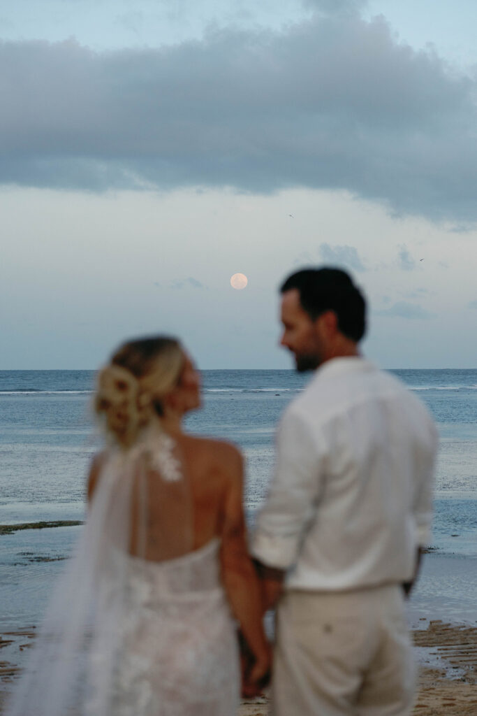Bride and groom holding hands on the beach at dusk in Bali, photographed out of focus against the ocean horizon and a rising full moon beneath dramatic evening clouds.