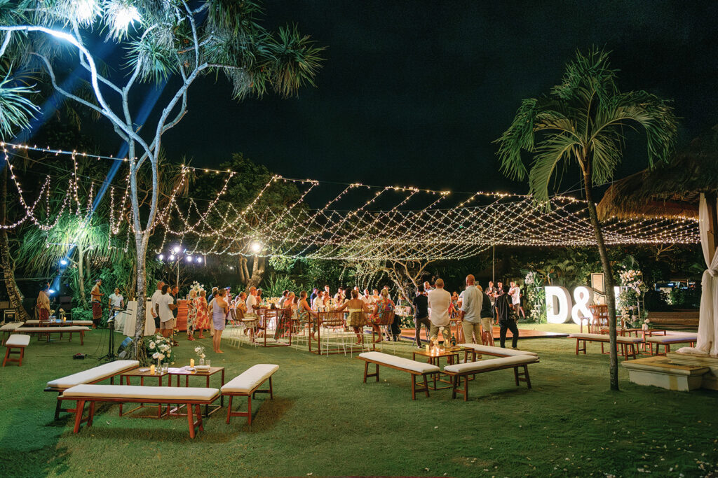 Warm outdoor wedding reception at Nusa Dua Beach Hotel Bali with fairy lights strung above the lawn, guests gathered around long tables, illuminated “D & J” monogram letters, tropical palm trees, and a relaxed nighttime celebration atmosphere.