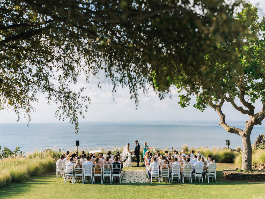 Clifftop wedding ceremony at Puri Bhagawan Bali overlooking the ocean with guests gathered beneath tropical trees at sunset.