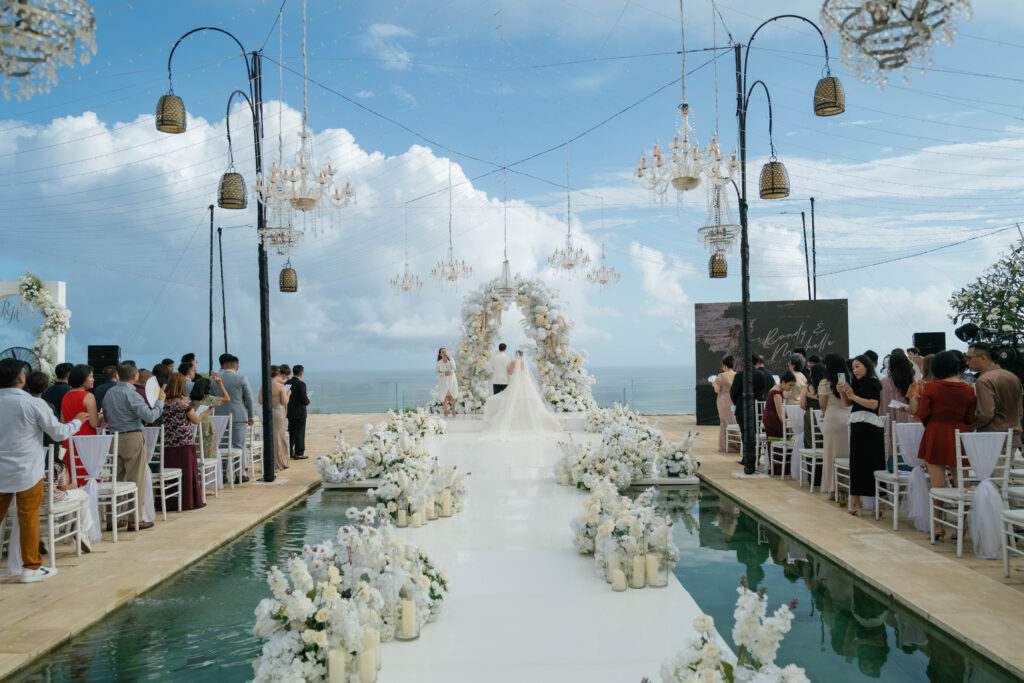 Villa Plenilunio Uluwatu infinity pool ceremony aisle with white floral arrangements hanging chandeliers and ocean backdrop — Luxima Wedding