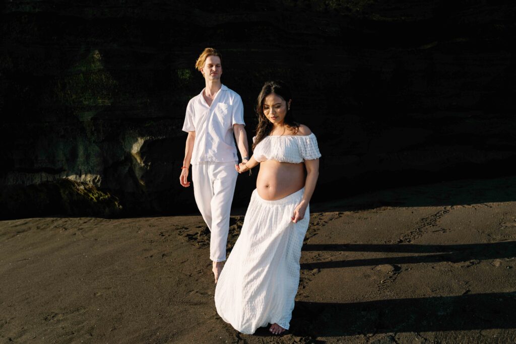 Elly and Tom walking on black sand at Nyanyi Beach Bali maternity session cliff backdrop