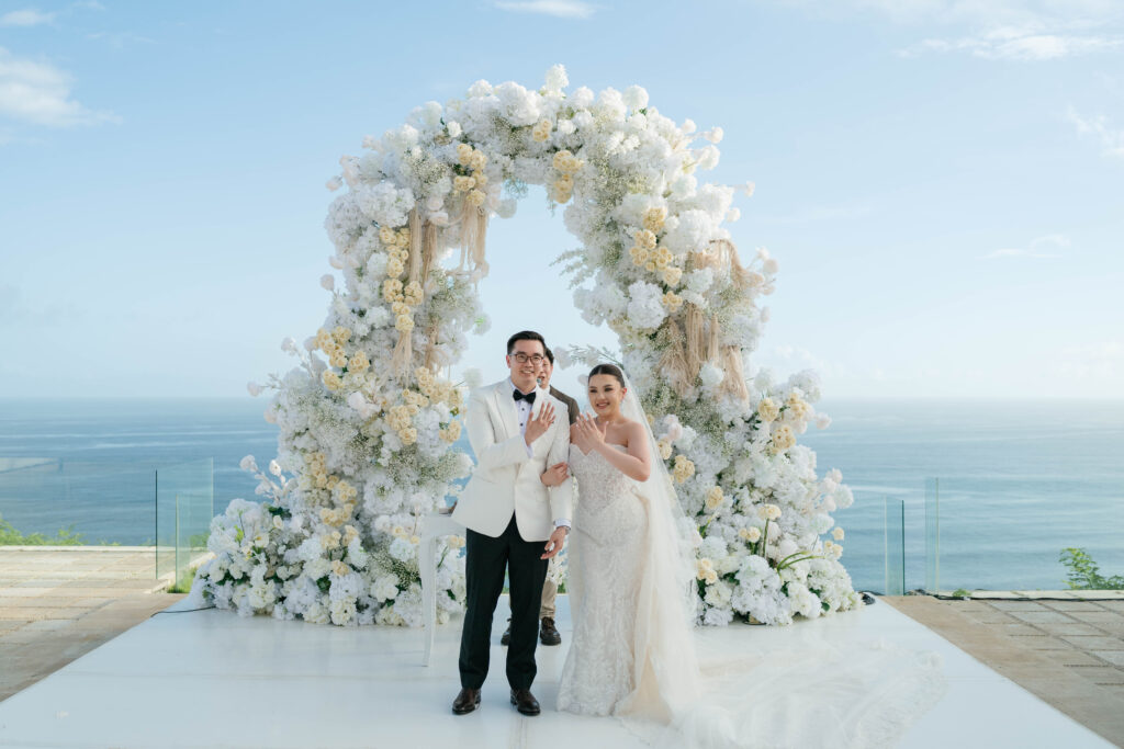 Randy and Michelle showing wedding rings in front of white floral arch at Villa Plenilunio Uluwatu with Indian Ocean behind — Luxima Wedding