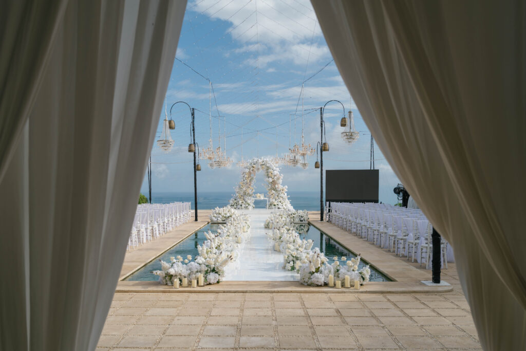 Villa Plenilunio Uluwatu wedding ceremony setup — white floral aisle along infinity pool with ocean horizon and hanging chandeliers — Luxima Wedding