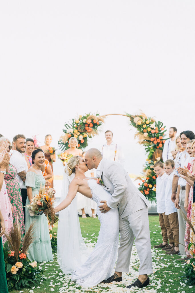 Bride and groom sharing their first kiss at a Bali wedding ceremony with tropical floral arch and guests celebrating
