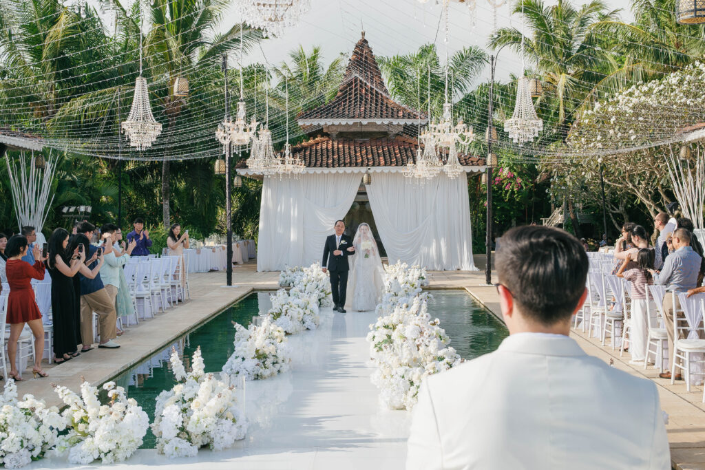 Michelle walking down Villa Plenilunio Uluwatu pool aisle with antique Javanese joglo structure in background during wedding processional — Luxima Wedding